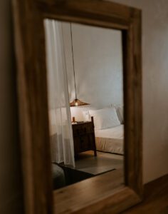 Un miroir en bois reflète une chambre lumineuse et paisible avec lit, voilage et lampe, symbolisant l’impact du placement des miroirs dans l’environnement